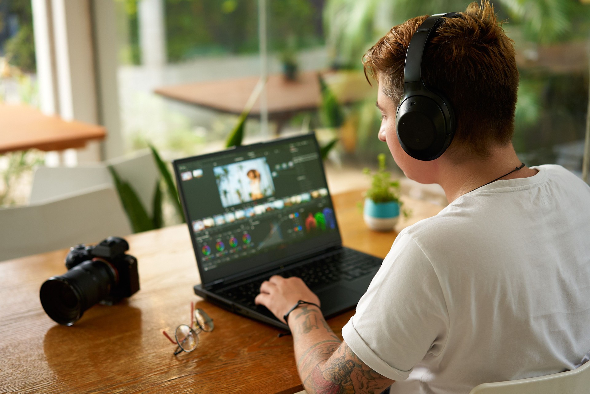 Professional video editor works on laptop in cozy cafe. Freelancer with tattoos wearing headphones edits film, digital camera on desk surrounded by plants by large window, daylight creative process.